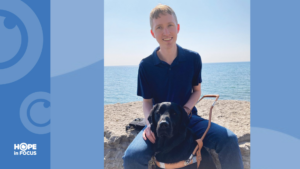 A young man sits on a stone ledge by the water with a guide dog resting between his legs